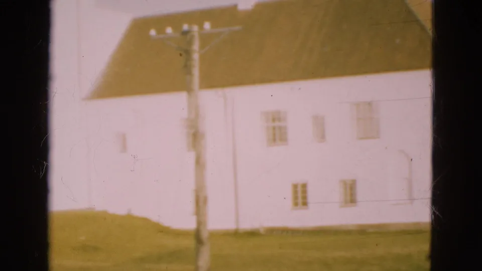1975 Denmark 8mm view of a pale rural house, grassy ground and utility pole framed by dark window bars.