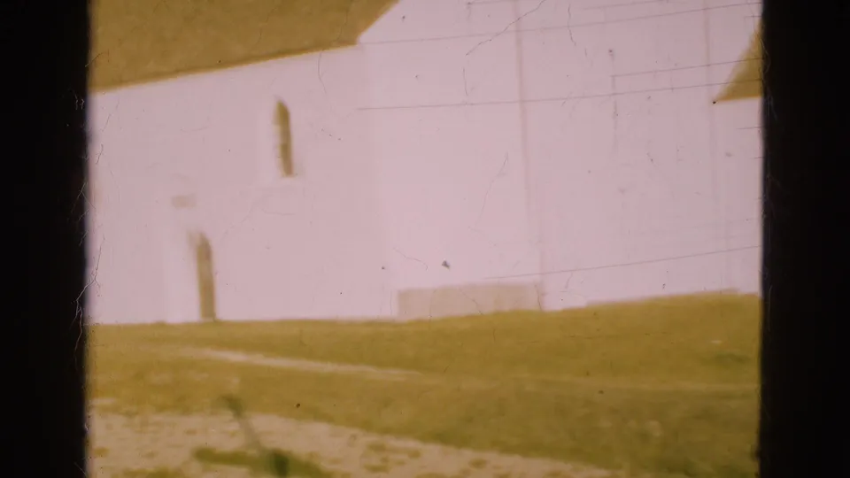 Blurred 1975 Denmark home-movie frame showing a white building corner, arched doorway, grass and window-frame shadow.