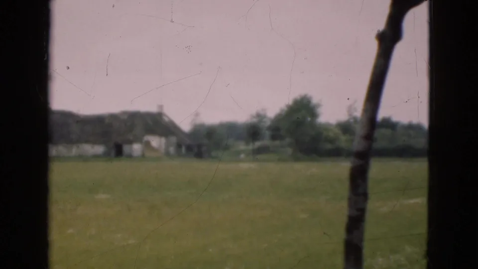 1975 Denmark 8mm view across a green field toward distant low buildings, bordered by dark window-frame edges.