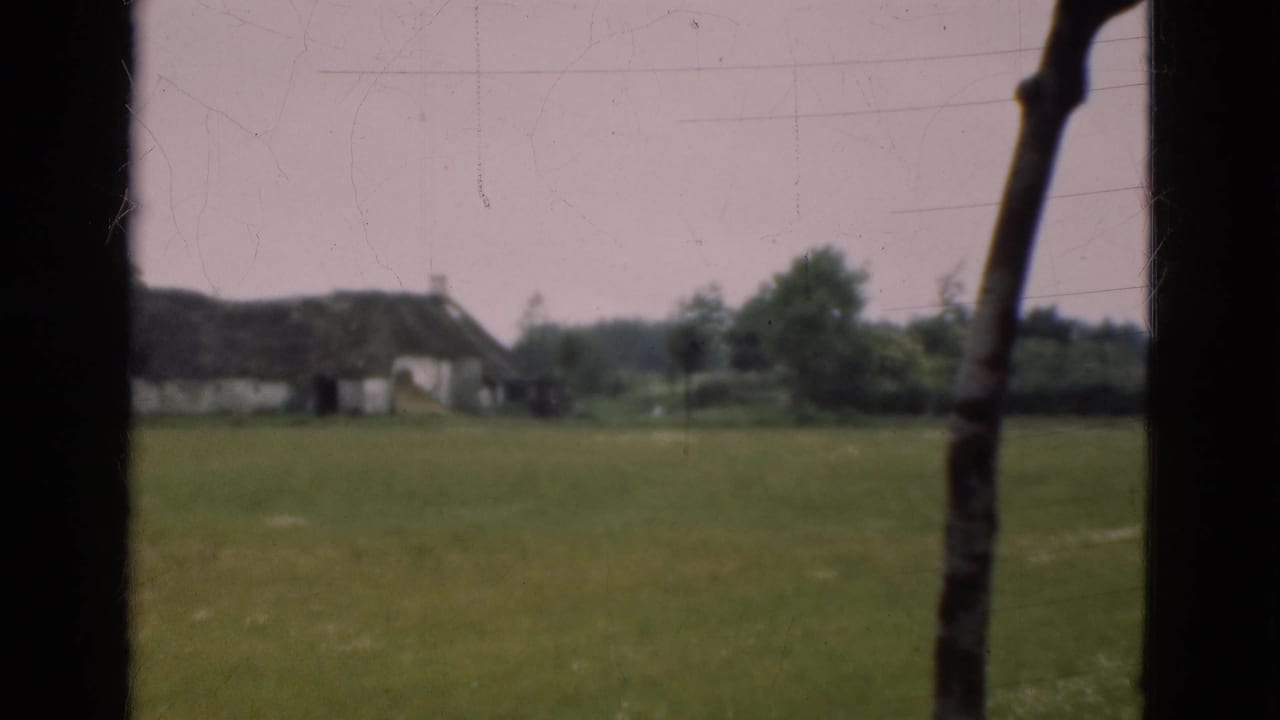 1975 Denmark home-movie view of a thatched cottage beyond a green pasture, framed by dark vertical window or doorway elements.