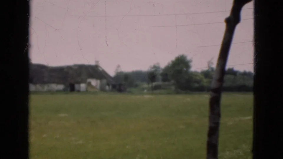 1975 Denmark home-movie view of a thatched cottage beyond a green pasture, framed by dark vertical window or doorway elements.