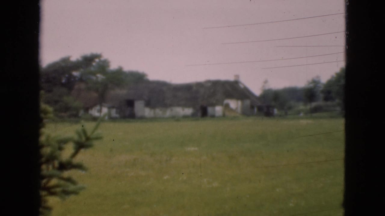Thatched rural buildings beyond a green field, seen through dark vertical framing.