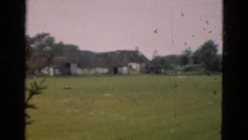 1975 Denmark frame of a grassy rural scene with distant thatched buildings, trees, pale sky, and visible 8mm surface marks.