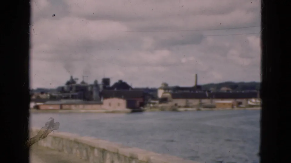 1975 Denmark home-movie view of a waterfront industrial area with low buildings, smokestacks, water, and a cloudy sky.