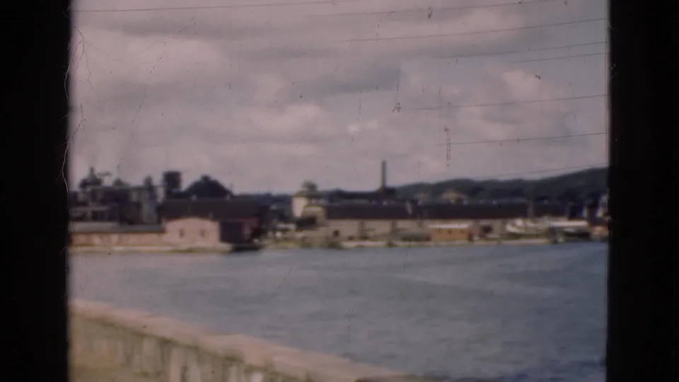 1975 Denmark frame showing water in the foreground and an industrial coastal shoreline with buildings and a smokestack under clouds.