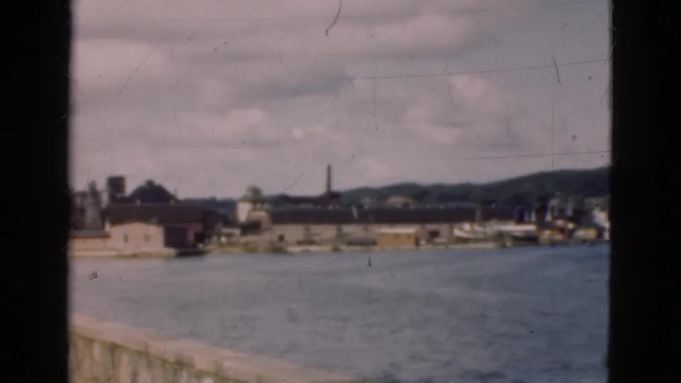 Danish harbor shoreline in 1975 seen from a moving vessel, with water, industrial buildings, a smokestack, hills, and window framing.