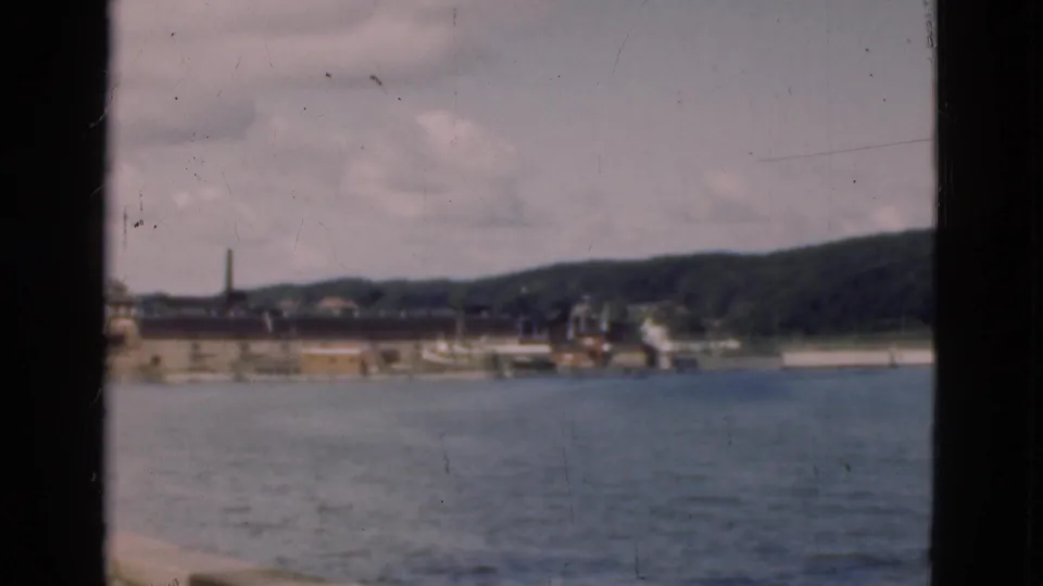Soft 8mm frame of a Danish harbor in 1975, showing water in the foreground, a pier or quay, industrial structures, a smokestack, and hills.