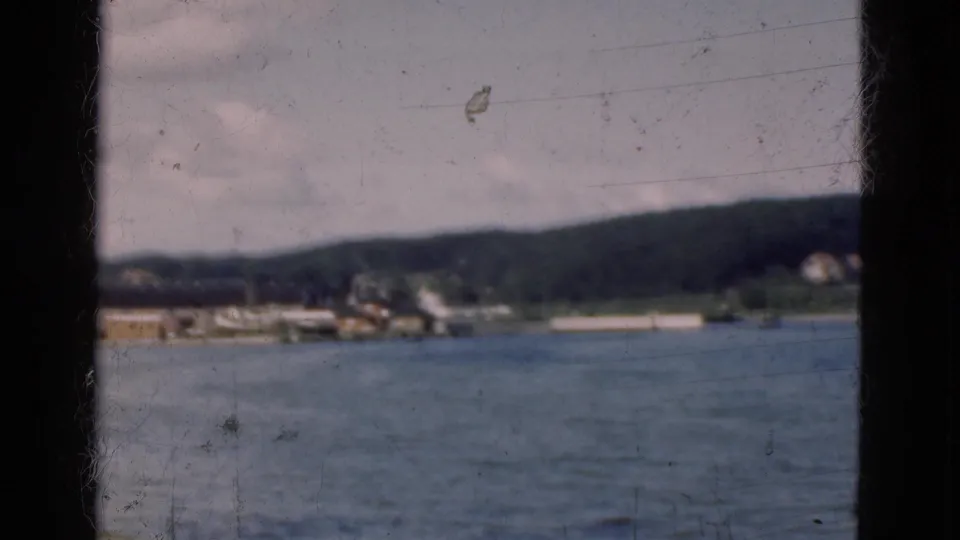 End frame of the 1975 Denmark port approach, with blue-gray water, low harbor structures, moored vessels, green hills, cloudy sky, and film wear.