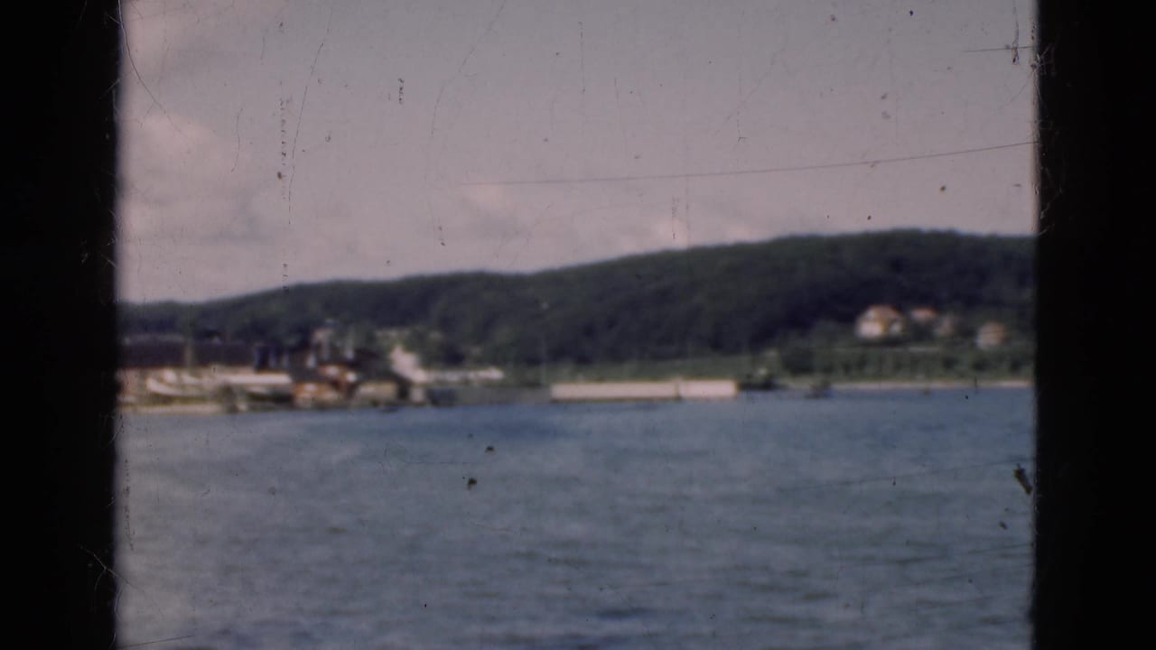 1975 Denmark home-movie view across water from a vessel, framed by a dark window edge with hills and shoreline buildings beyond.