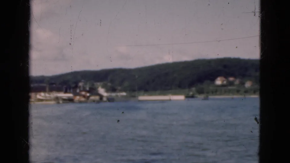 1975 Denmark home-movie view across water from a vessel, framed by a dark window edge with hills and shoreline buildings beyond.