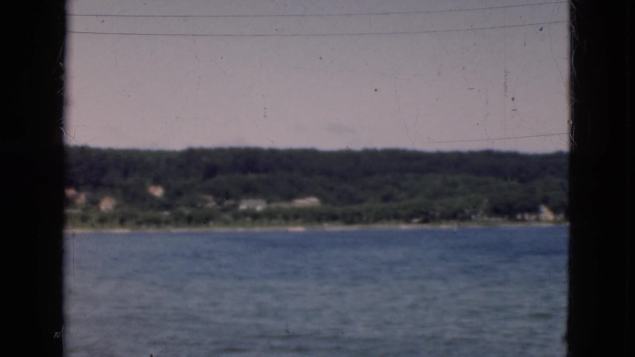 Soft 8mm frame of a Danish shoreline across calm water, with a dark window divider cutting through the boat-view composition.
