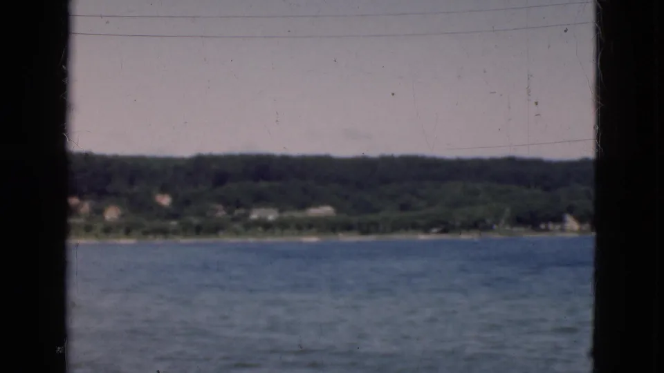 Soft 8mm frame of a Danish shoreline across calm water, with a dark window divider cutting through the boat-view composition.
