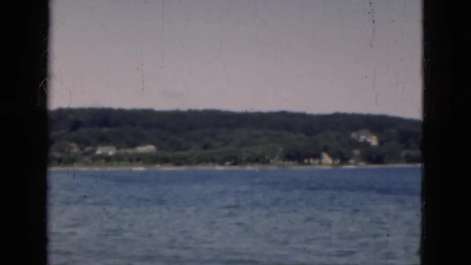 Vintage 8mm coastal view in Denmark, showing rippled water, a low green shore, and distant buildings under a pale sky.