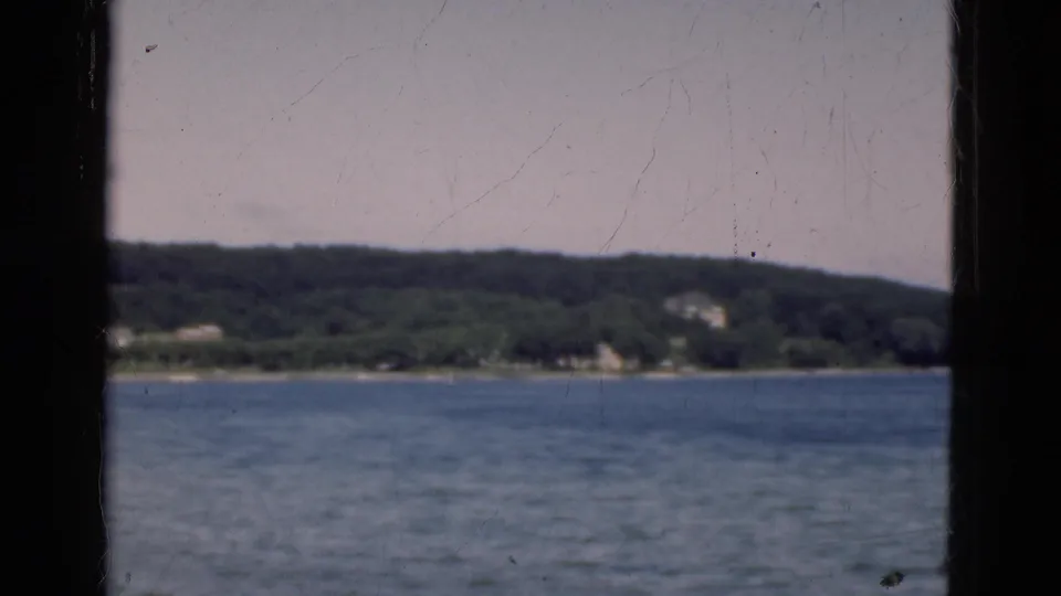 End-frame view across Danish coastal water toward trees, scattered shore buildings, and a wooded hillside in soft 8mm focus.