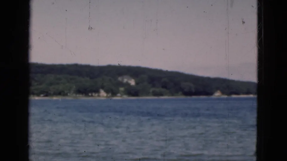 1975 Denmark 8mm frame with calm lake water, forested shore, small distant structures, and a dark vertical divider in the foreground.