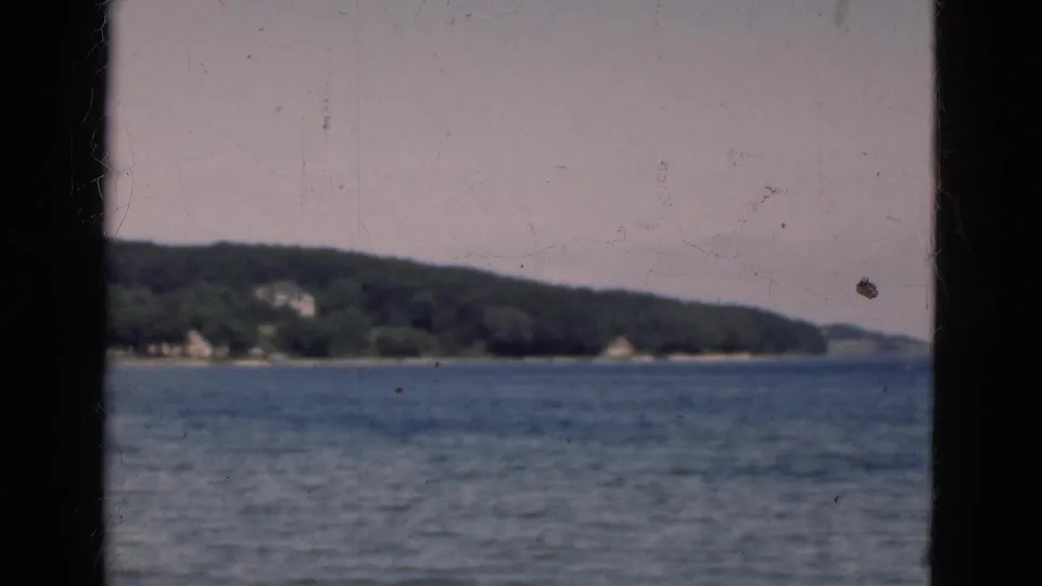 1975 Denmark 8mm frame showing a broad lake view, low forested shoreline, distant light structures, and visible film scratches.