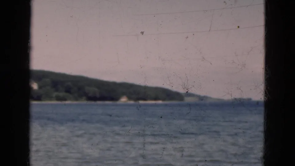 1975 Denmark 8mm frame with a soft lake horizon, tree-lined shore, faint distant buildings, and dark framing from a nearby structure.