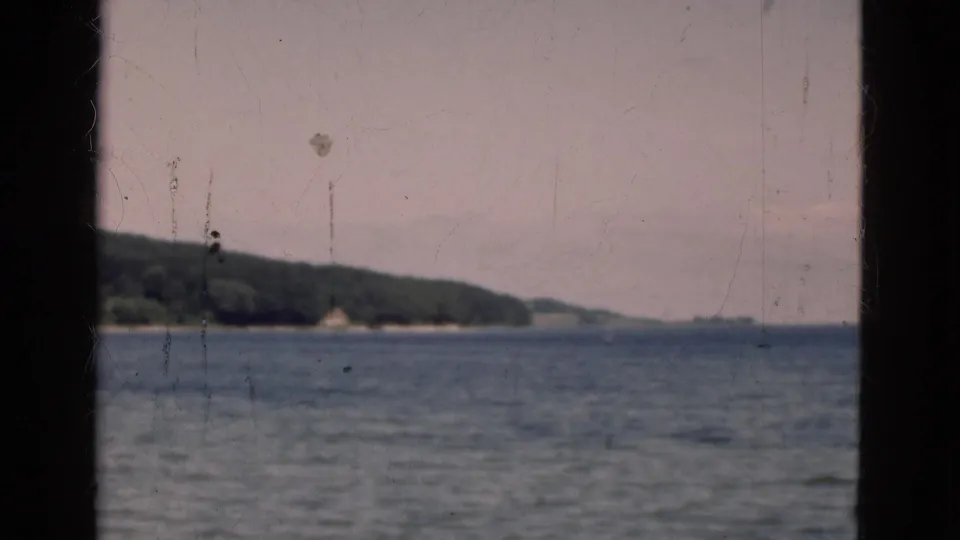 1975 Denmark 8mm frame showing open water, a wooded shore, and distant low hills from a moving viewpoint.
