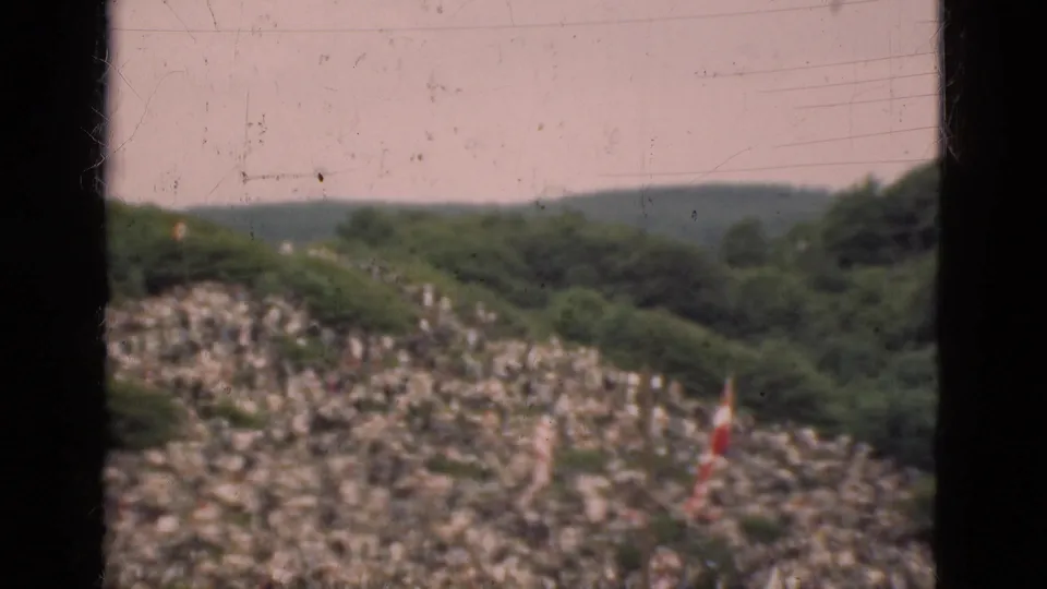 1975 Denmark 8mm frame showing distant spectators on a hillside near dense trees and a red-and-white vertical object.
