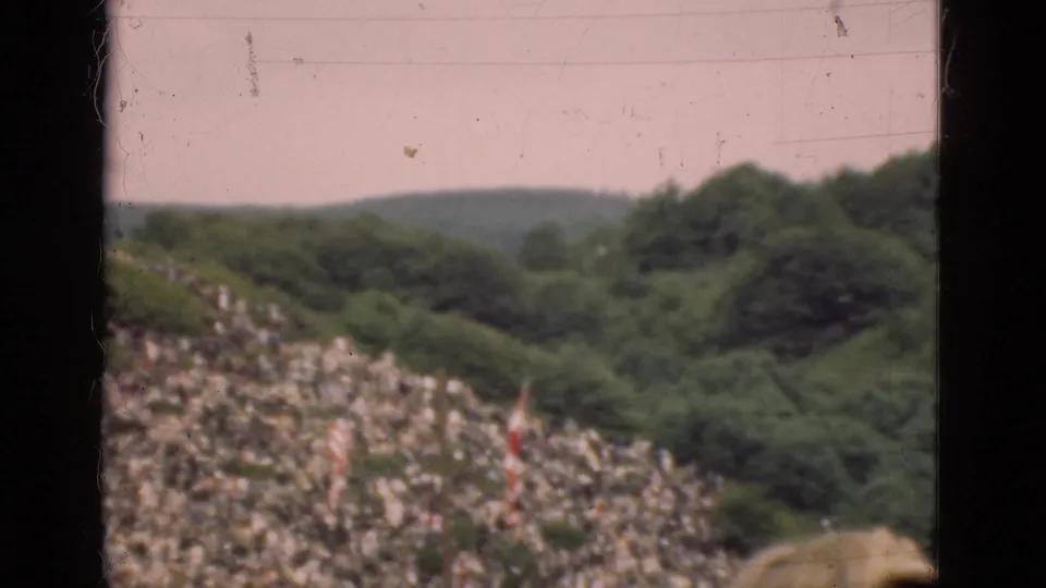 1975 Denmark 8mm frame showing a wooded slope with distant people, low hills, and a partial foreground obstruction.