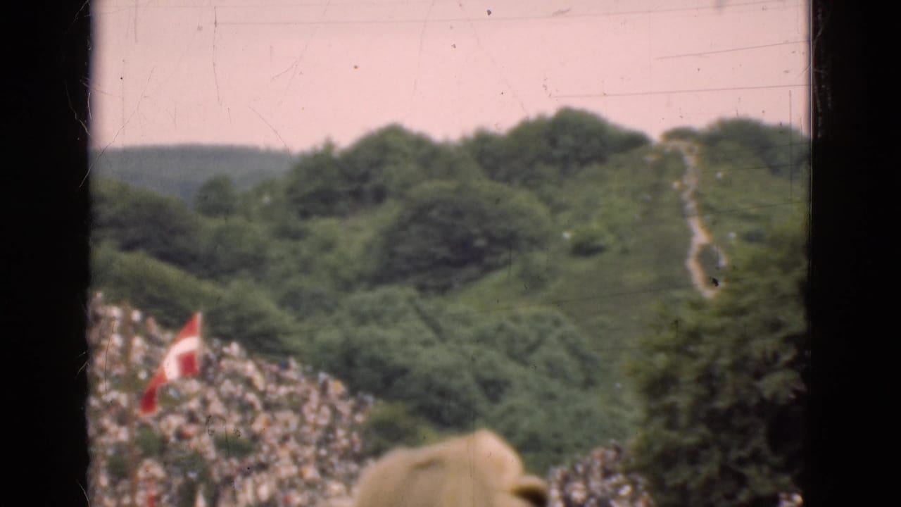 1975 Denmark home-movie frame with a Danish flag, crowded slope, trees, and a pale hillside path in the background.