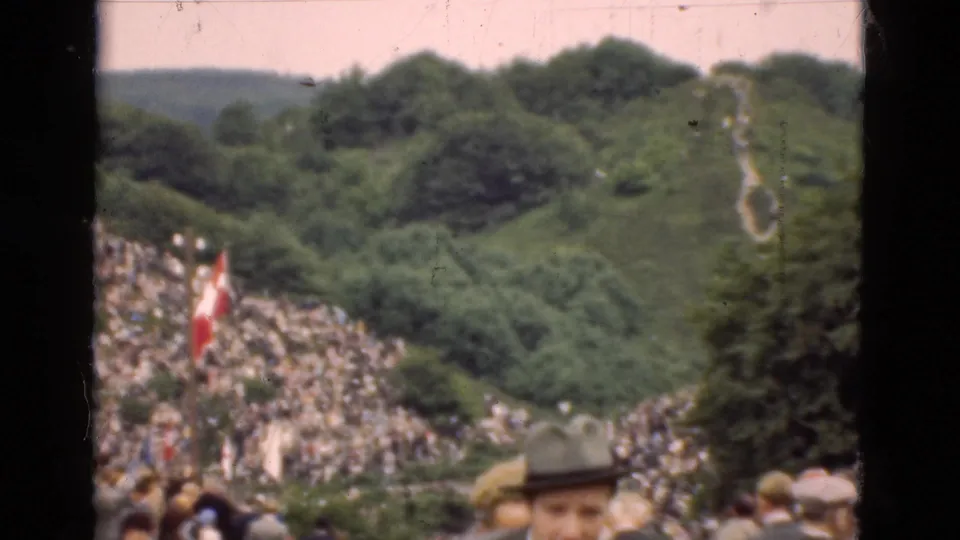 1975 Denmark 8mm view of a public hillside crowd, Danish flag, dense trees, and foreground spectators in hats.