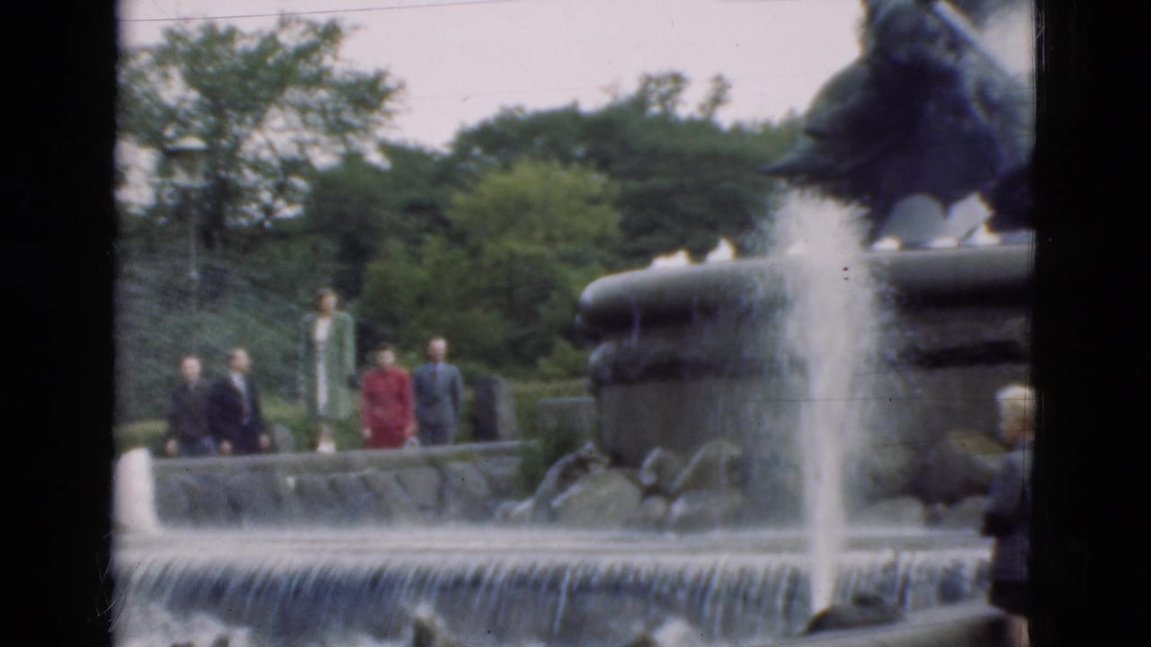 In Denmark 1975 home-movie footage, visitors in suits and dresses stand near a cascading fountain with a dark sculptural figure above it.