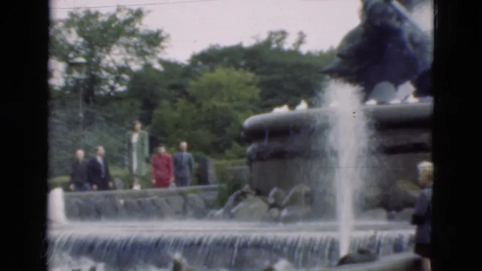 In Denmark 1975 home-movie footage, visitors in suits and dresses stand near a cascading fountain with a dark sculptural figure above it.