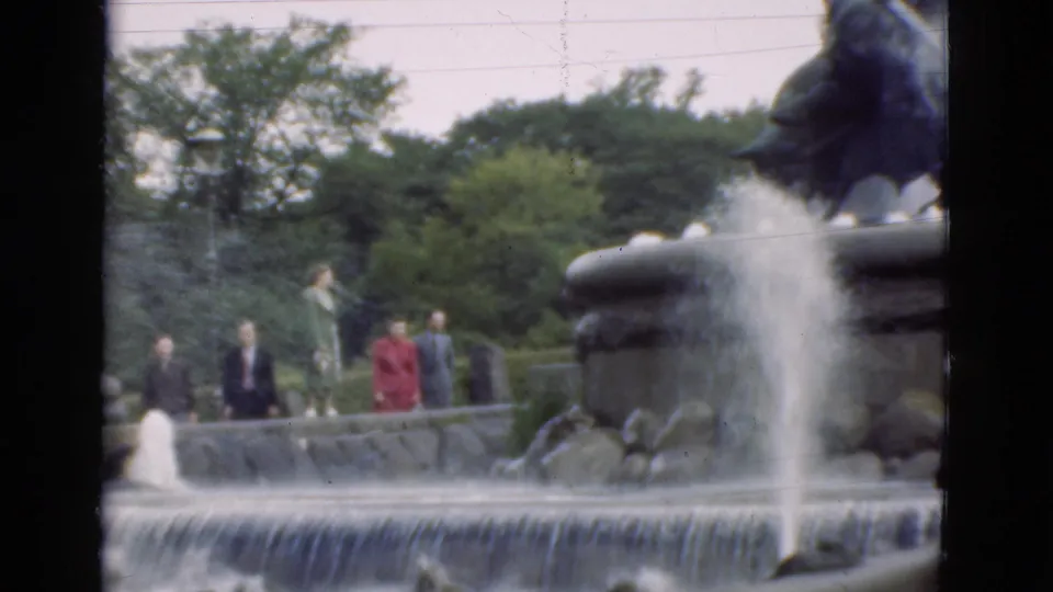 Visitors in period clothing stand by a Danish park fountain, with water spraying near a dark stone sculptural element and trees in the background.