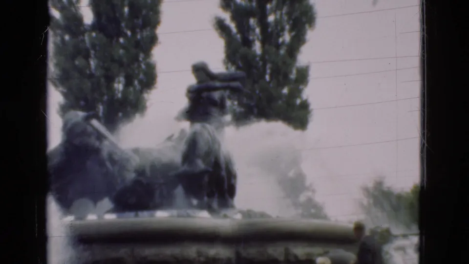 A closer home-movie view shows the fountain's dark sculptural figures, water spray, stone rim, and tall trees in the background.