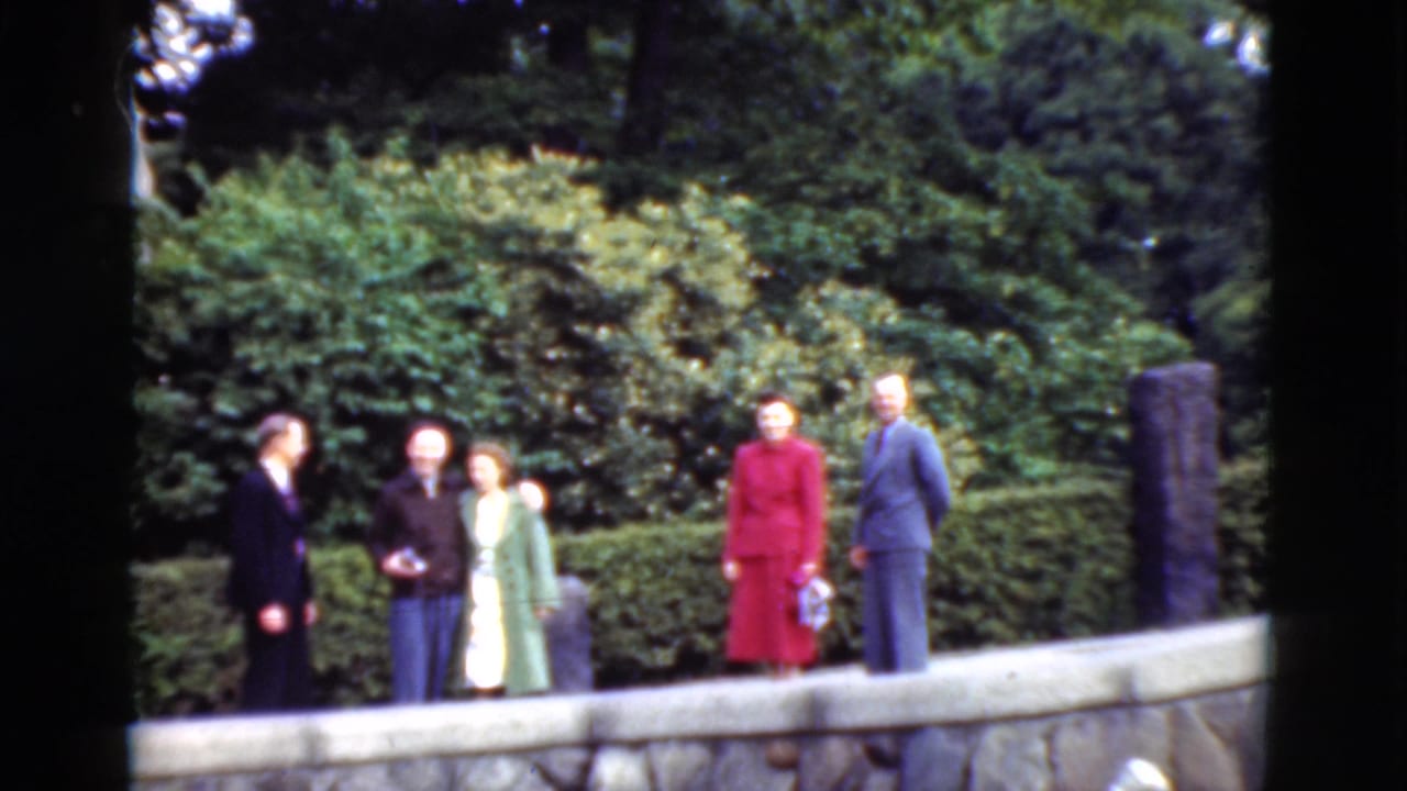 In metadata-supported 1975 Denmark footage, adults in suits and dresses gather beside a stone wall in a landscaped outdoor setting.