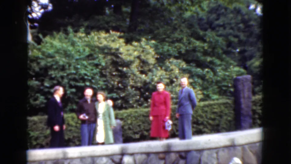 In metadata-supported 1975 Denmark footage, adults in suits and dresses gather beside a stone wall in a landscaped outdoor setting.