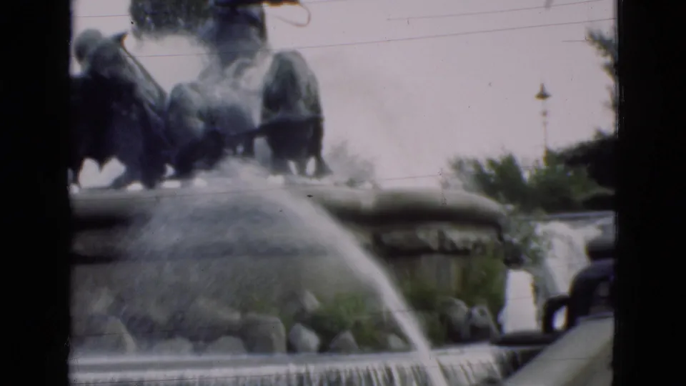 1975 Denmark 8mm frame showing a tiered public fountain with cascading water, rocks, greenery, and dark sculptures at the top.