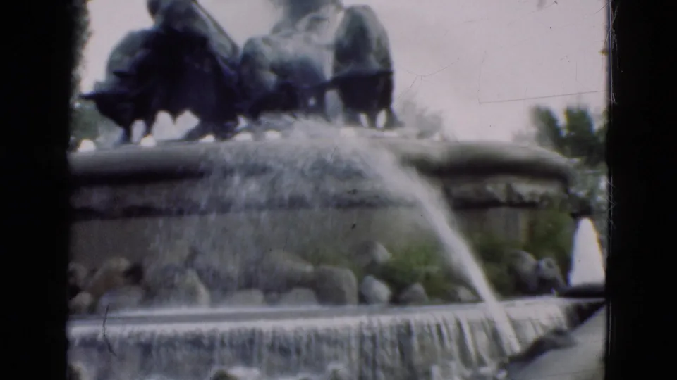 1975 Denmark home-movie frame of a sculptural park fountain with flowing water, stone tiers, rocks, shrubs, and pale sky.