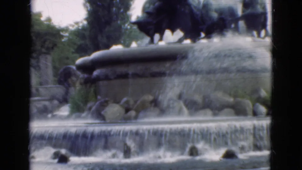 1975 Denmark visitor-shot 8mm view of a broad cascading fountain, rocky lower basin, shrubs, trees, and dark sculpture overhead.
