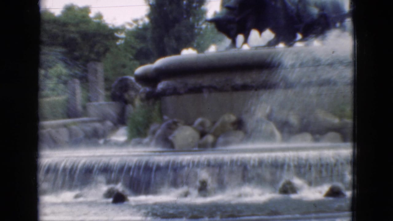 Fountain water cascades below a dark statue, partly divided by black film frame bars.