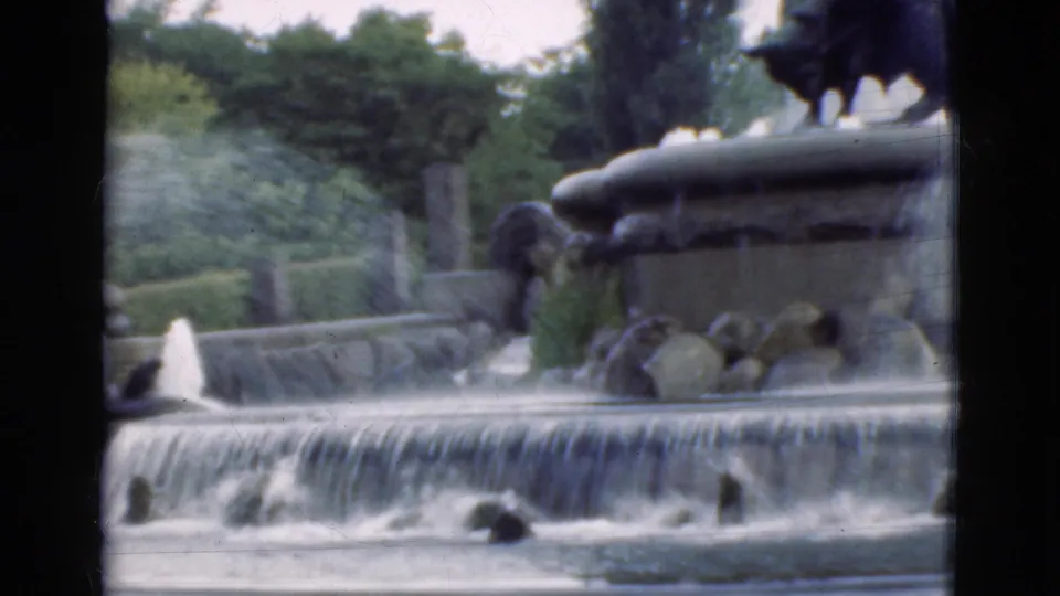 1975 Denmark 8mm frame of a public park fountain, showing water spray, a stone basin, greenery, and a dark statue cropped at the top.