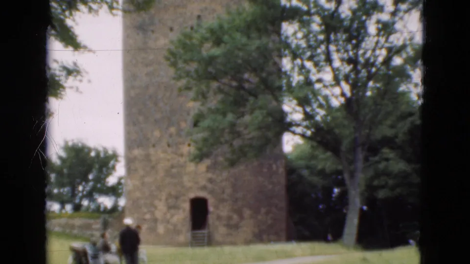 1975 Denmark 8mm frame showing a stone tower in a park with trees, lawn, benches, distant visitors, and dark film or window-frame bars.