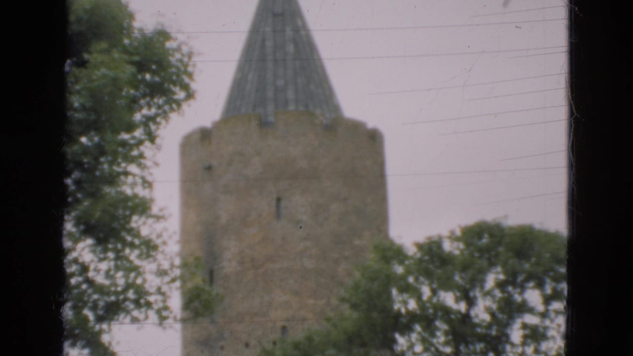 1975 Denmark 8mm frame showing a tall stone tower with a conical spire behind leafy trees under a pale sky.