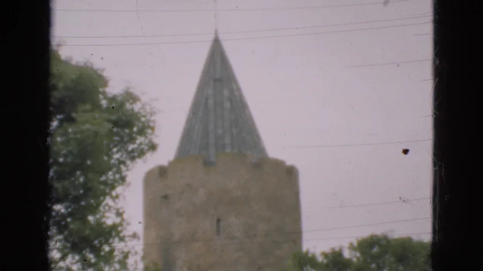 1975 Denmark frame of a round stone tower and steep spire, partly obscured by trees and film gate edges.