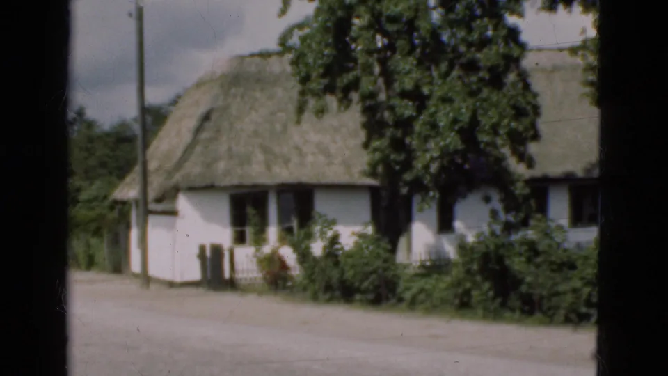 1975 Denmark 8mm frame showing a white thatched cottage near a roadside, with trees and garden shrubs visible.