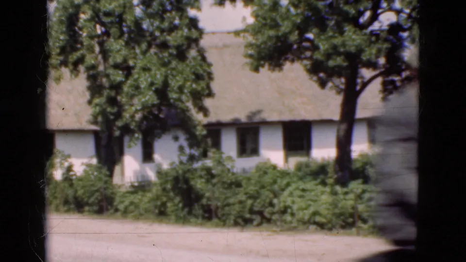 A man on a bicycle moves past the roadside cottage in this 1975 Denmark home-movie frame, blurred by motion.