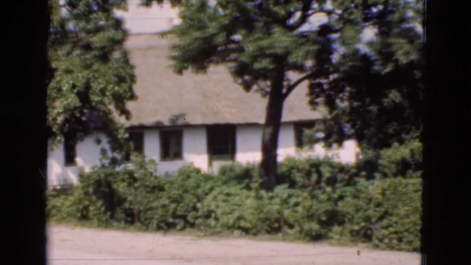 1975 Denmark home-movie view of a thatched cottage and roadside garden framed by trees, with no cyclist visible in this still.