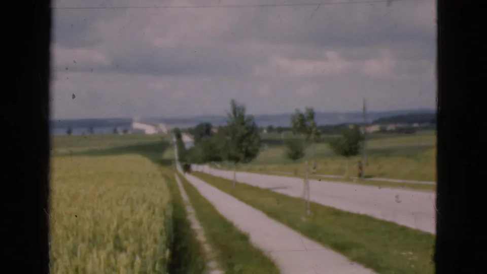 1975 Danish home-movie frame of open fields and a rural roadside, with small trees and low buildings in the distance.