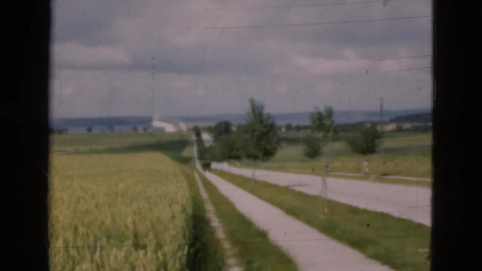 1975 Denmark 8mm frame showing a narrow rural road between fields, with scattered trees and distant farm structures seen through dark window edges.