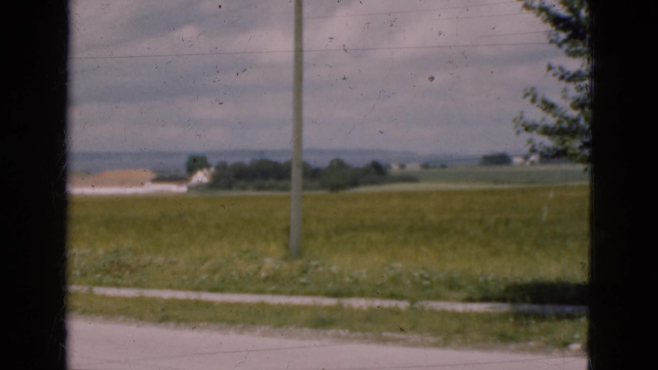 Open fields with a utility pole, distant low buildings, and dark vertical window framing.