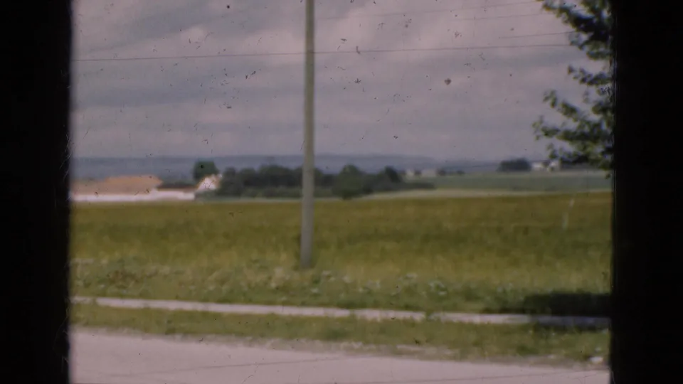 1975 Denmark countryside view with grassy fields, a roadside utility pole, distant farm buildings, and dark window bars crossing the frame.