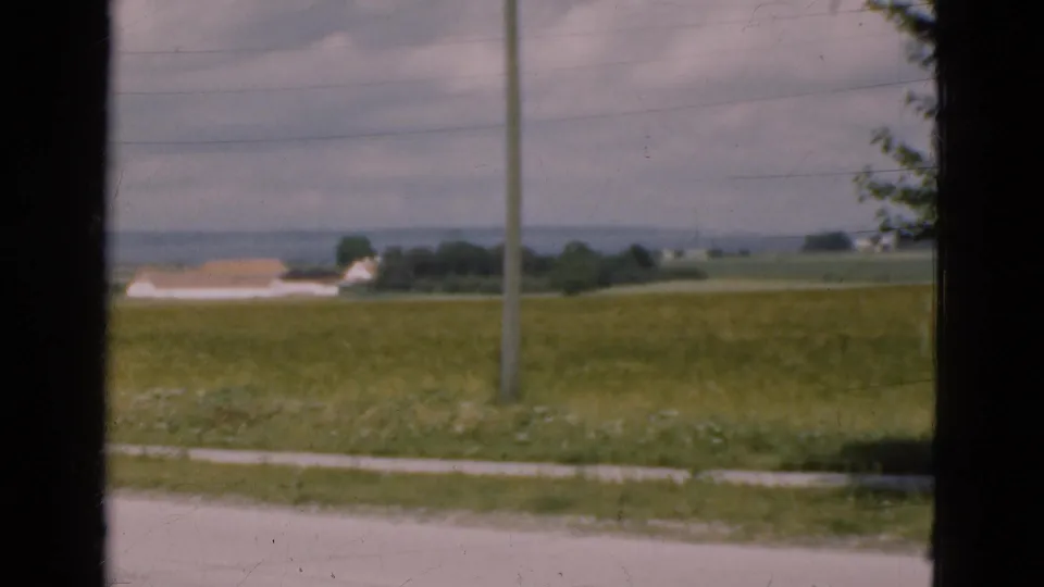 1975 Denmark rural frame showing fields, a utility pole, distant farm buildings, and a pale roadside edge viewed from a moving vehicle or window.