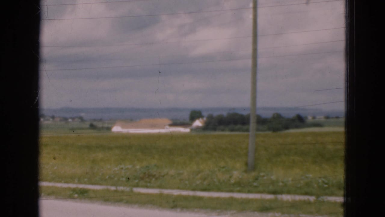 Green roadside field with a distant pale building, utility pole, cloudy sky, and dark window frame.