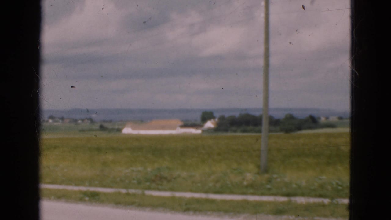 Rural field seen through dark window framing, with utility pole, distant building, and cloudy sky.